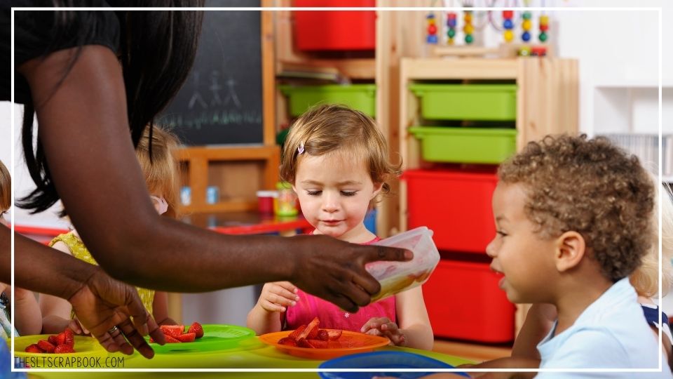Picture of a group of children sat around a table. An adult is offering each child a few pieces of strawberry. A visual example of the 'communication temptations' early language strategy.